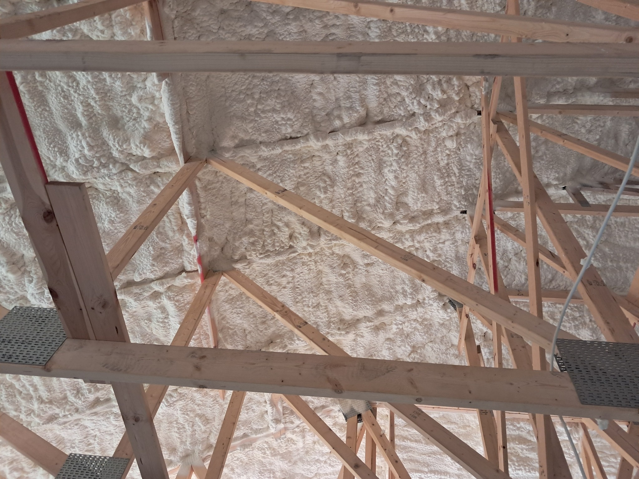 Poddasze z drewnianą konstrukcją pokryte grubą warstwą białej piany izolacyjnej, widoczne metalowe elementy mocujące.