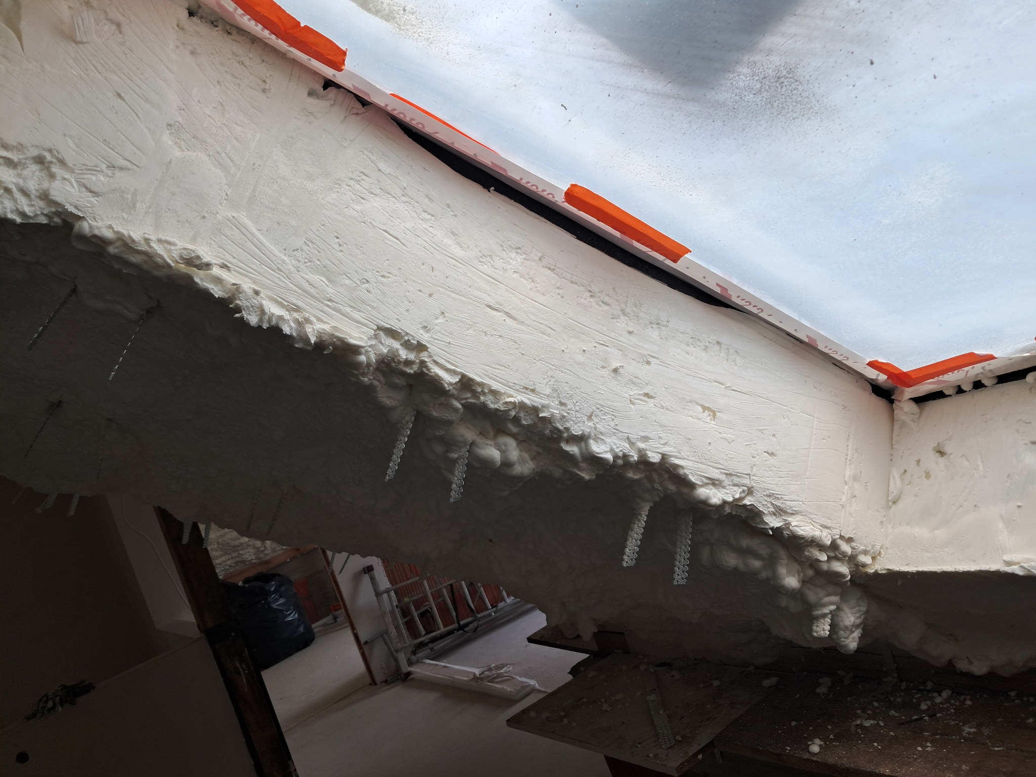 Wnętrze poddasza w trakcie prac izolacyjnych, widoczna biała pianka montażowa wypełniająca przestrzeń między krokwiami, z widocznymi kotwami mocującymi i fragmentami folii paroizolacyjnej...