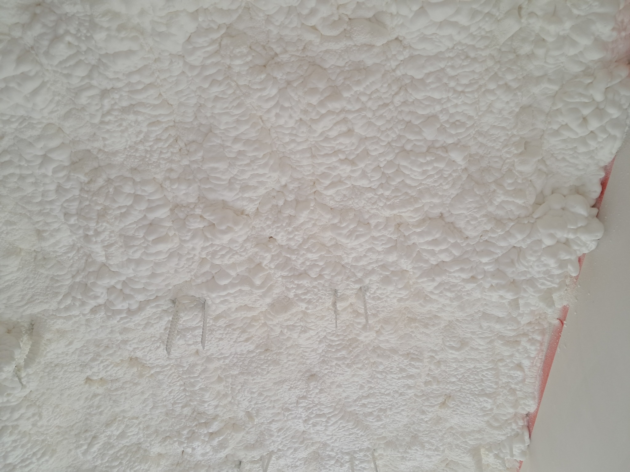 Pianka izolacyjna o strukturze popcornu, przytwierdzona do poddasza za pomocą metalowych zszywek; widoczne fragmenty konstrukcji dachu.