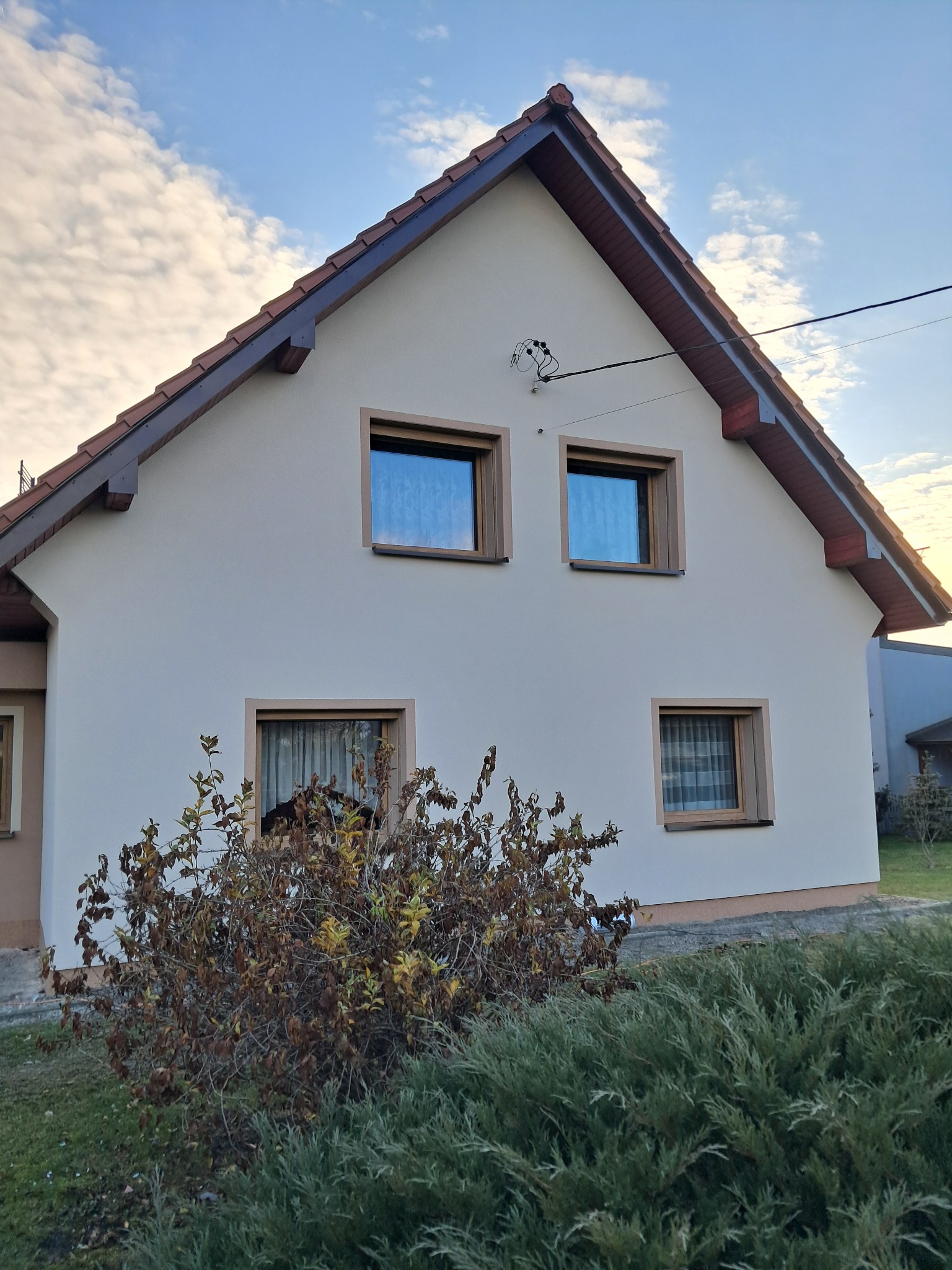 Dom jednorodzinny z jasną elewacją, brązowymi ramami okiennymi i dachem z czerwonej dachówki, widoczny krzew na pierwszym planie.