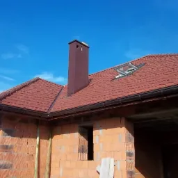 Niedokończony dom z czerwonej cegły z zamontowanym dachem z dachówki ceramicznej, brązowy komin i okno dachowe, widoczne błękitne niebo.