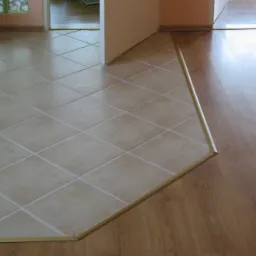 Połączenie beżowych płytek podłogowych ułożonych w karo z panelami podłogowymi w kolorze jasnego drewna, oddzielone listwą progową.