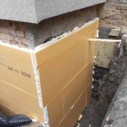 Ż&oacute;łte płyty izolacyjne XPS, marki BACHL, przyklejone pianką do fundamentu budynku, widoczne wykopane obok fundament&oacute;w podłoże i fragment folii kubełkowej.