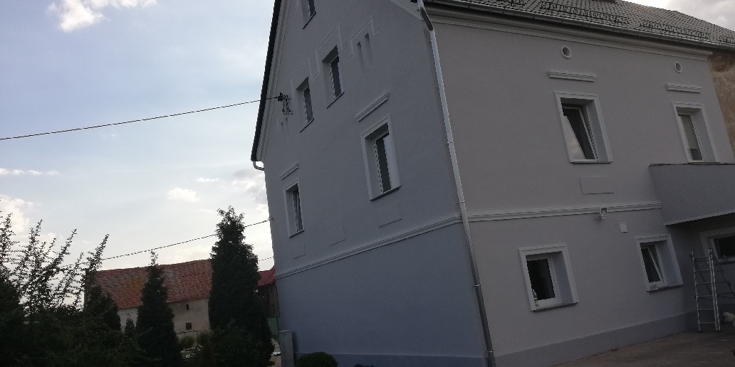 Odnowiona elewacja dwukondygnacyjnego budynku z dekoracyjnymi elementami wokół okien, pomalowana na szaro z ciemniejszym odcieniem na dolnej części ściany, widoczna z poziomu gruntu z fragmentem...