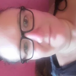 Portret kobiety w okularach z ciemnymi oprawkami na różowym tle. Ujęcie z bliska, włosy upięte, ciemna bluzka. Zdjęcie obrócone o 90 stopni.