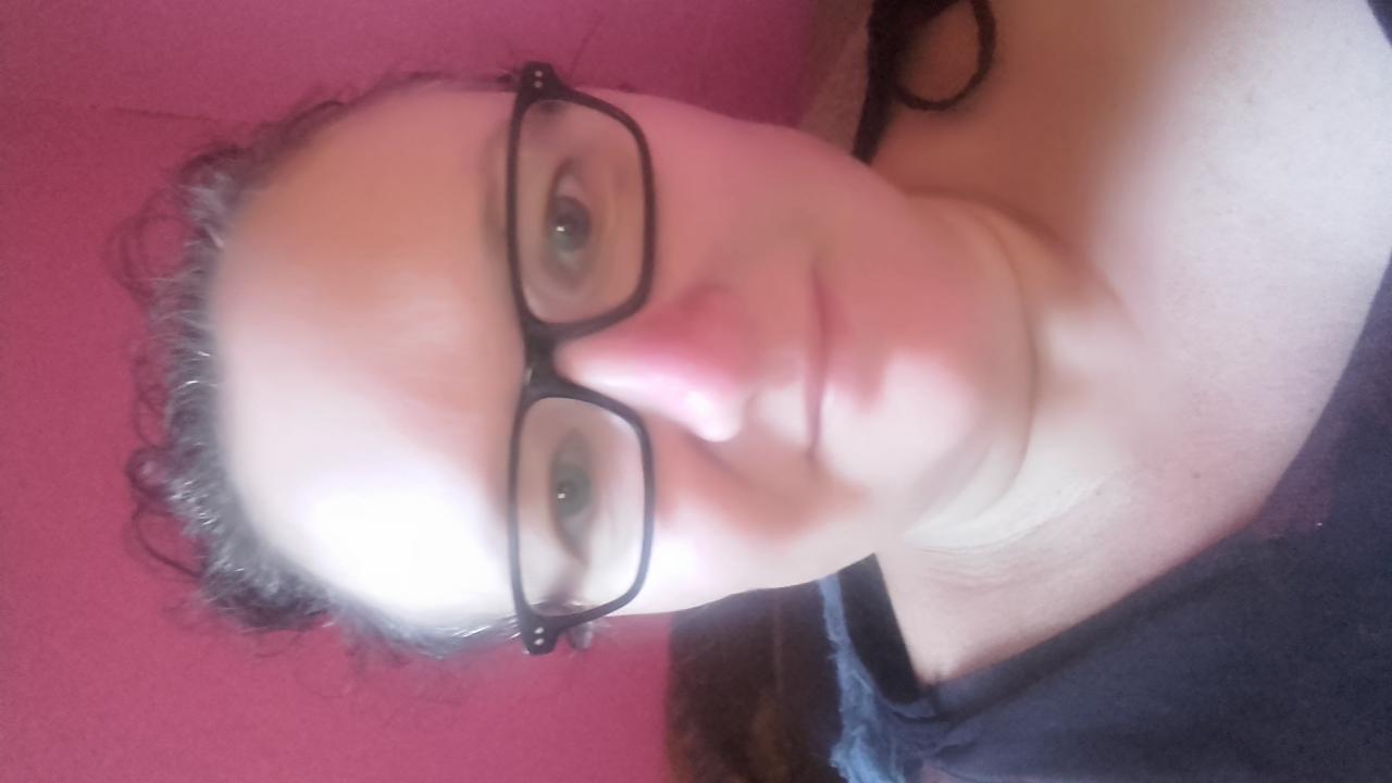 Portret kobiety w okularach z ciemnymi oprawkami na różowym tle. Ujęcie z bliska, włosy upięte, ciemna bluzka. Zdjęcie obrócone o 90 stopni.