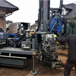 Operator obsługuje wiertnicę na gąsienicach z niebieską obudową i napisem 'Kreto XXX', obok stoi mężczyzna w okularach, w tle budowa domu i zaparkowany biały van.