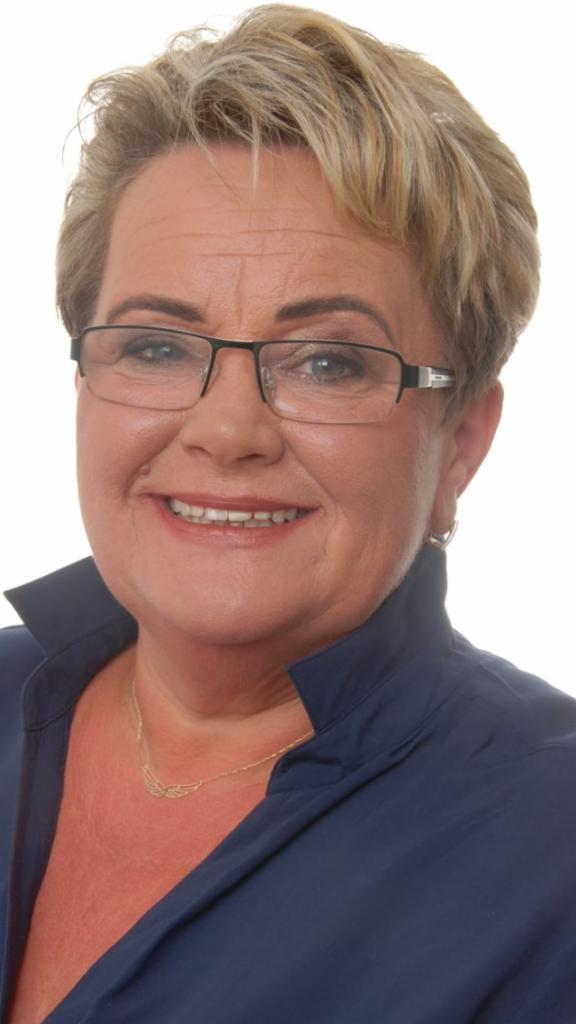 Portret kobiety w okularach, z krótkimi blond włosami, ubrana w granatową koszulę. Widoczny złoty łańcuszek. Na białym tle.