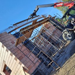 R Klawe Home firma budowlana Robert Klawe - Czerwony podnośnik teleskopowy Manitou umieszcza prefabrykowane elementy zbrojenia na wznoszonej konstrukcji budynku z pustaków ceramicznych, widoczne szalunki i zbrojenie.