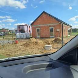 R Klawe Home firma budowlana Robert Klawe - Budowa domu z czerwonej cegły w trakcie realizacji. Widoczne okna, sterta worków z zaprawą, piasek i fragment deski rozdzielczej samochodu.