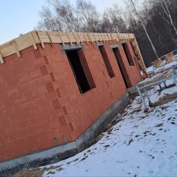 R Klawe Home firma budowlana Robert Klawe - Ściana budynku w trakcie wznoszenia z czerwonej cegły dziurawki, z widoczną konstrukcją drewnianą u góry. Zimowy krajobraz z białym śniegiem.