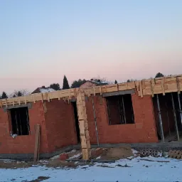 Wznoszenie ścian domu z czerwonej cegły Porotherm. Widoczne drewniane szalunki nad otworami okiennymi i podporami. Zimowy krajobraz w tle, paleta z cegłami Wienerberger.