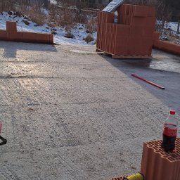 R Klawe Home firma budowlana Robert Klawe - Świeżo wylana płyta fundamentowa z paletą cegieł ceramicznych i butelką napoju. Widoczne narzędzia murarskie i zabudowa w tle. Słoneczny dzień.