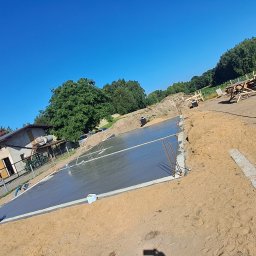 R Klawe Home firma budowlana Robert Klawe - Świeżo wylana, gładka płyta fundamentowa na budowie w słoneczny dzień. Widoczna zbrojenia, otoczenie placu budowy, częściowo dom i drzewa w tle.