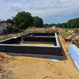 R Klawe Home firma budowlana Robert Klawe - Przygotowany zarys fundamentów budynku z hydroizolacją i ociepleniem, widok z góry na wykop z piaskiem, budynek gospodarczy i samochody w tle.