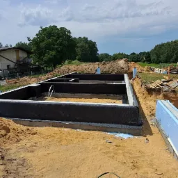 Przygotowany zarys fundamentów budynku z hydroizolacją i ociepleniem, widok z góry na wykop z piaskiem, budynek gospodarczy i samochody w tle.