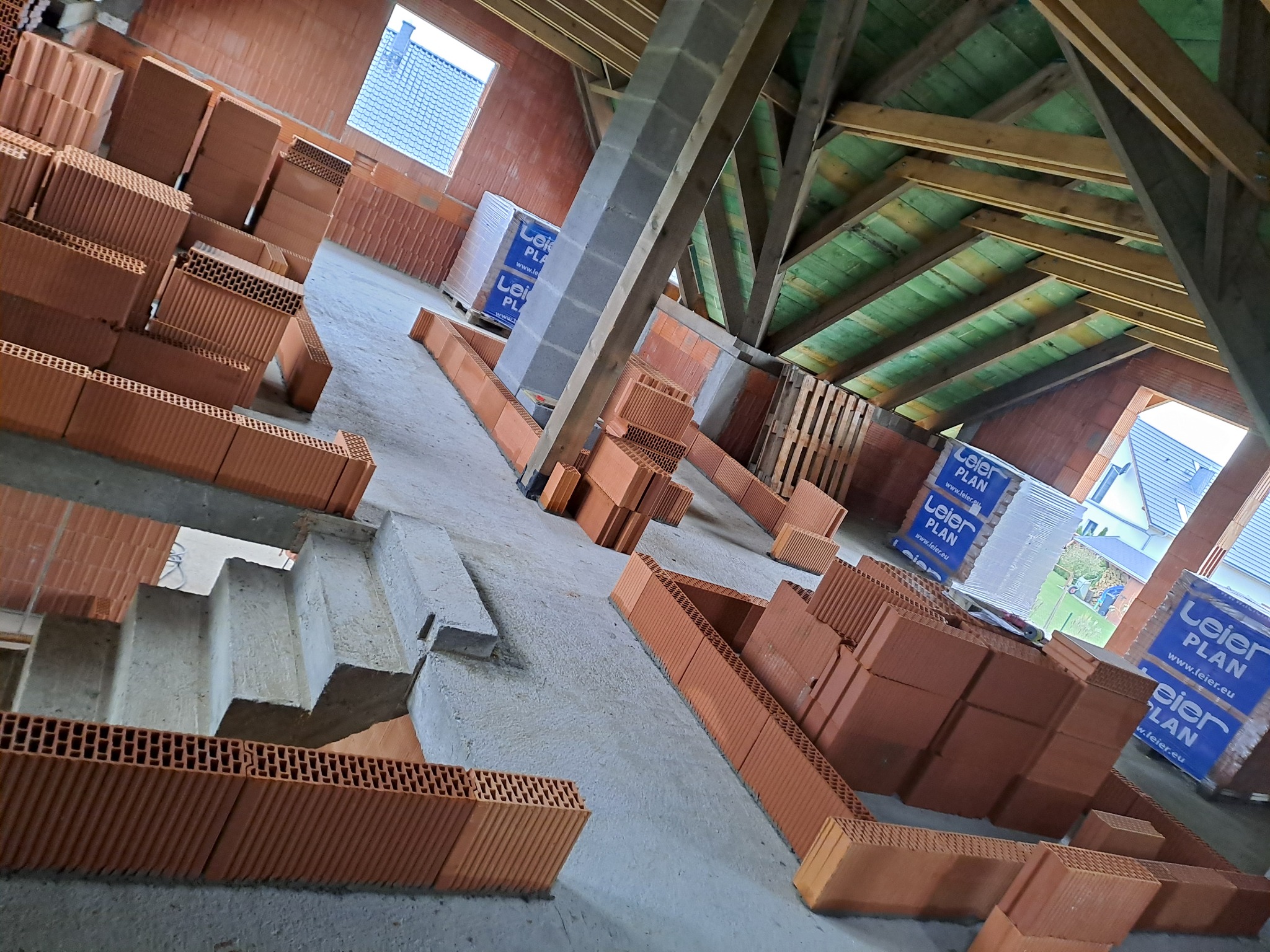 Wnętrze w trakcie budowy domu z cegły ceramicznej, widoczne ściany wznoszone z pustaków, drewniana konstrukcja dachu z zieloną izolacją, palety z materiałem budowlanym w tle, betonowe schody.