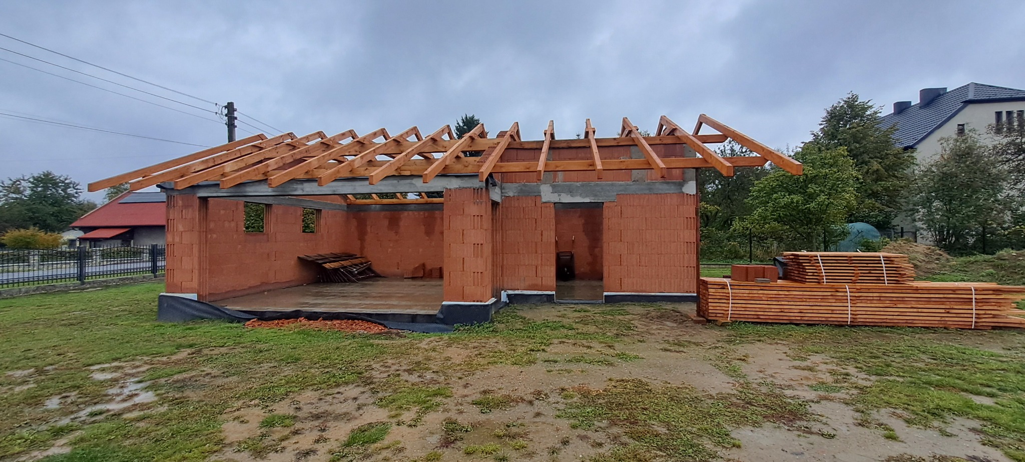 Surowy stan budynku w trakcie budowy z czerwonej cegły z drewnianą konstrukcją dachu, widok z zewnątrz na tle pochmurnego nieba i zieleni.