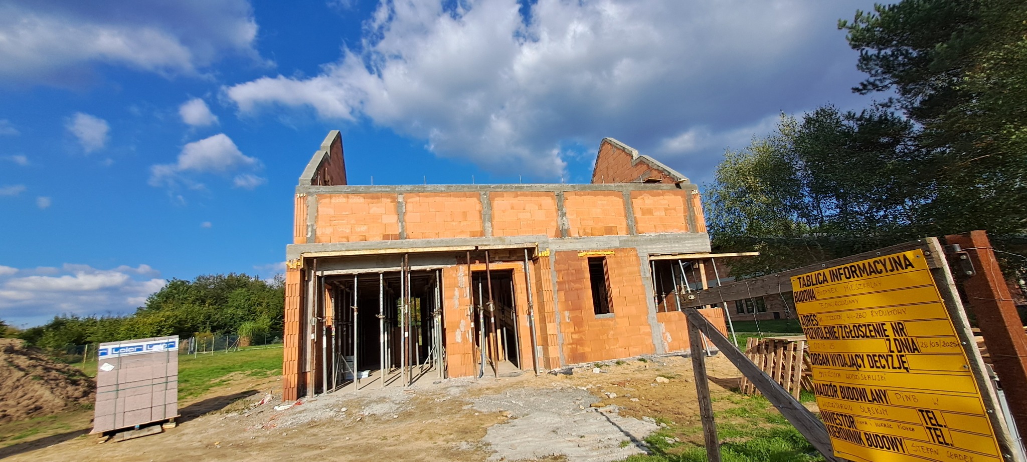 Budowa domu jednorodzinnego w trakcie murowania ścian. Widoczna tablica informacyjna budowy. Ściany z czerwonej cegły, niebo z chmurami w tle.
