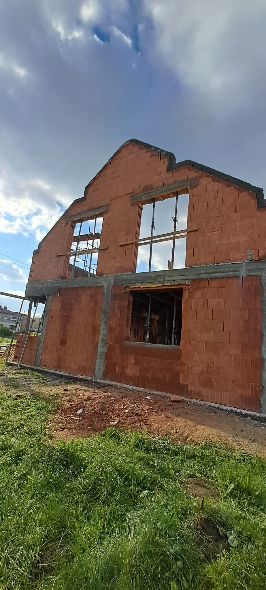 Wznoszony dwukondygnacyjny budynek z czerwonej cegły, z otworami okiennymi zabezpieczonymi stemplami budowlanymi, na tle błękitnego nieba z chmurami.