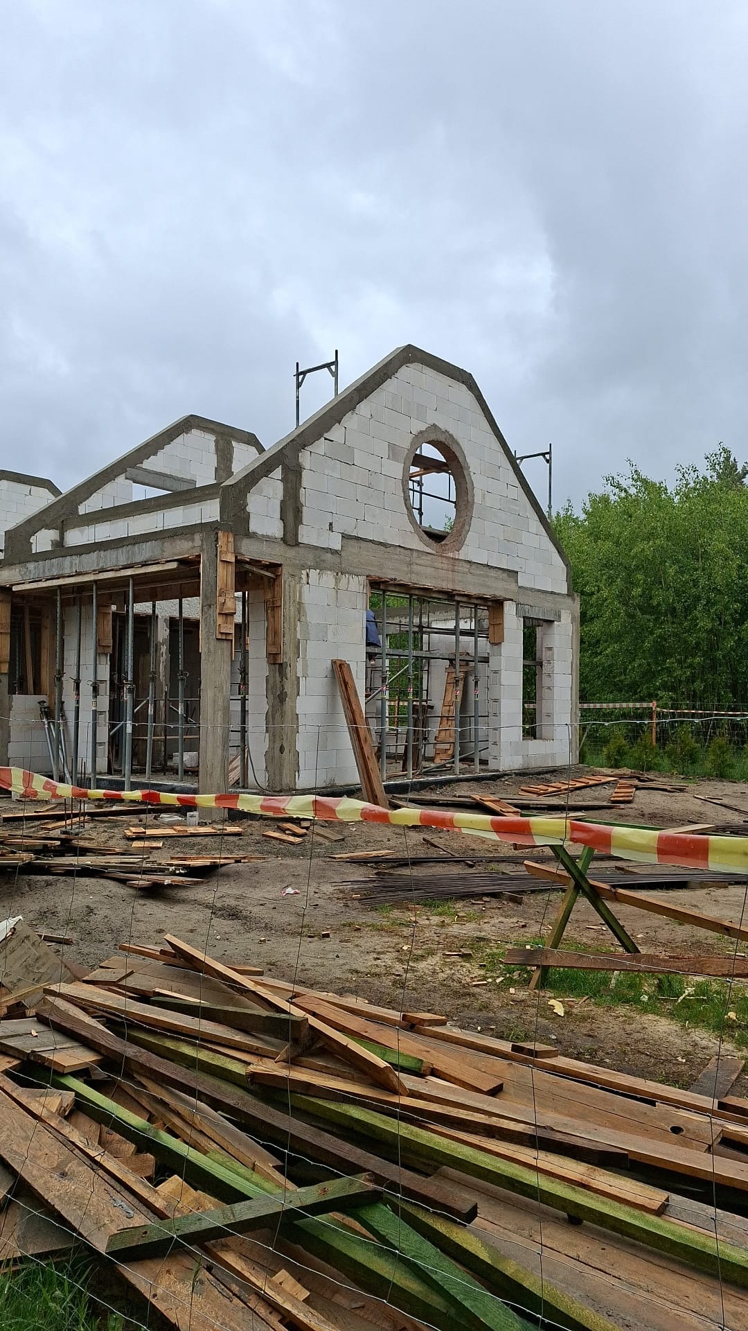 Budowa domu z białych bloczków z okrągłym oknem na szczycie. Widoczne drewniane podpory, teren budowy zabezpieczony taśmą, składowane deski. Etap murowania ścian.