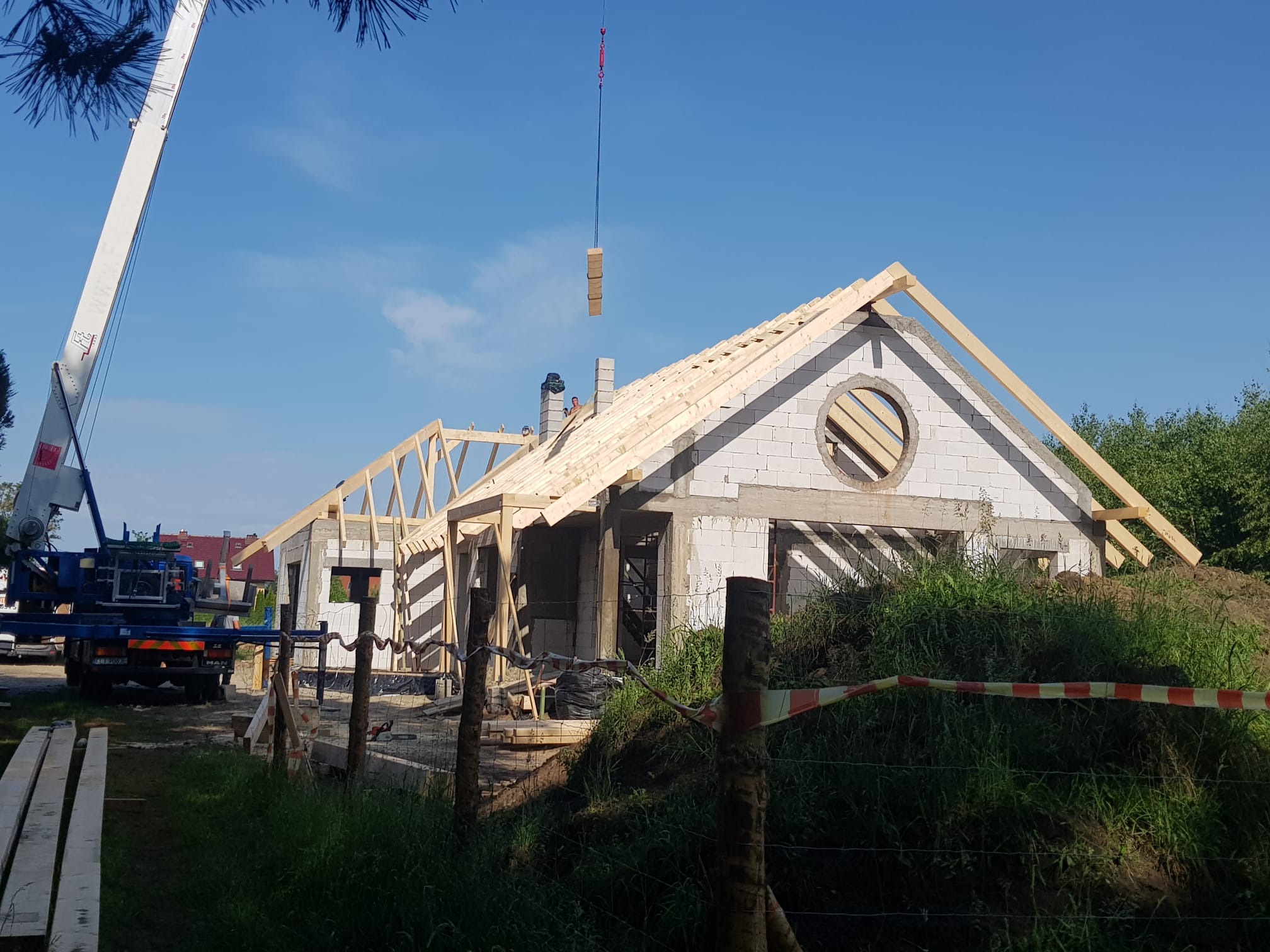 Budowa domu z białych bloczków, widoczny dźwig podający materiały na dach. Konstrukcja dachu w trakcie montażu. Okrągłe okno w szczycie. Teren budowy zabezpieczony.