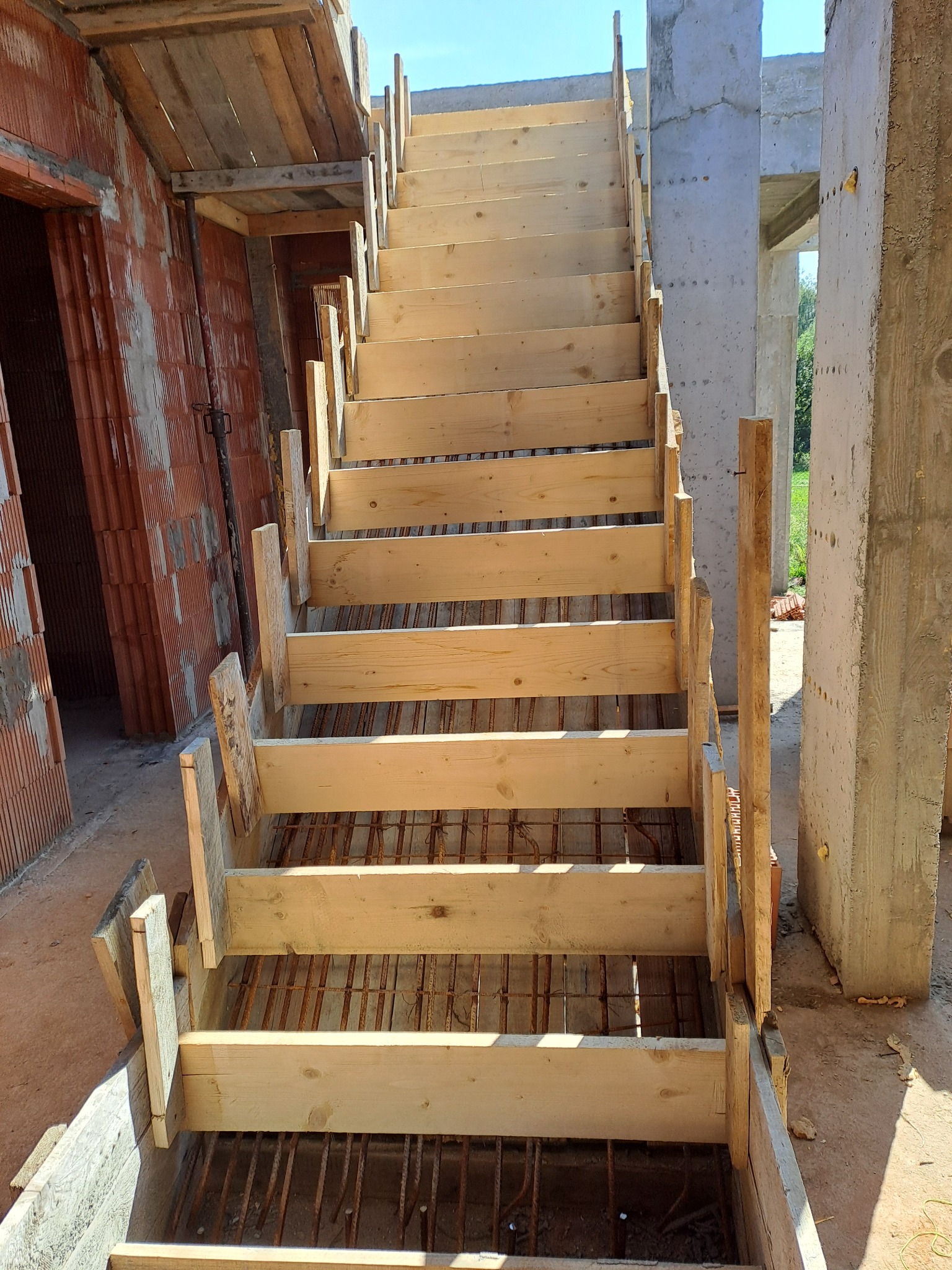Szczegółowe ujęcie drewnianego szalunku schodów z widocznym zbrojeniem, na tle surowych murów i betonowych kolumn w trakcie budowy domu.