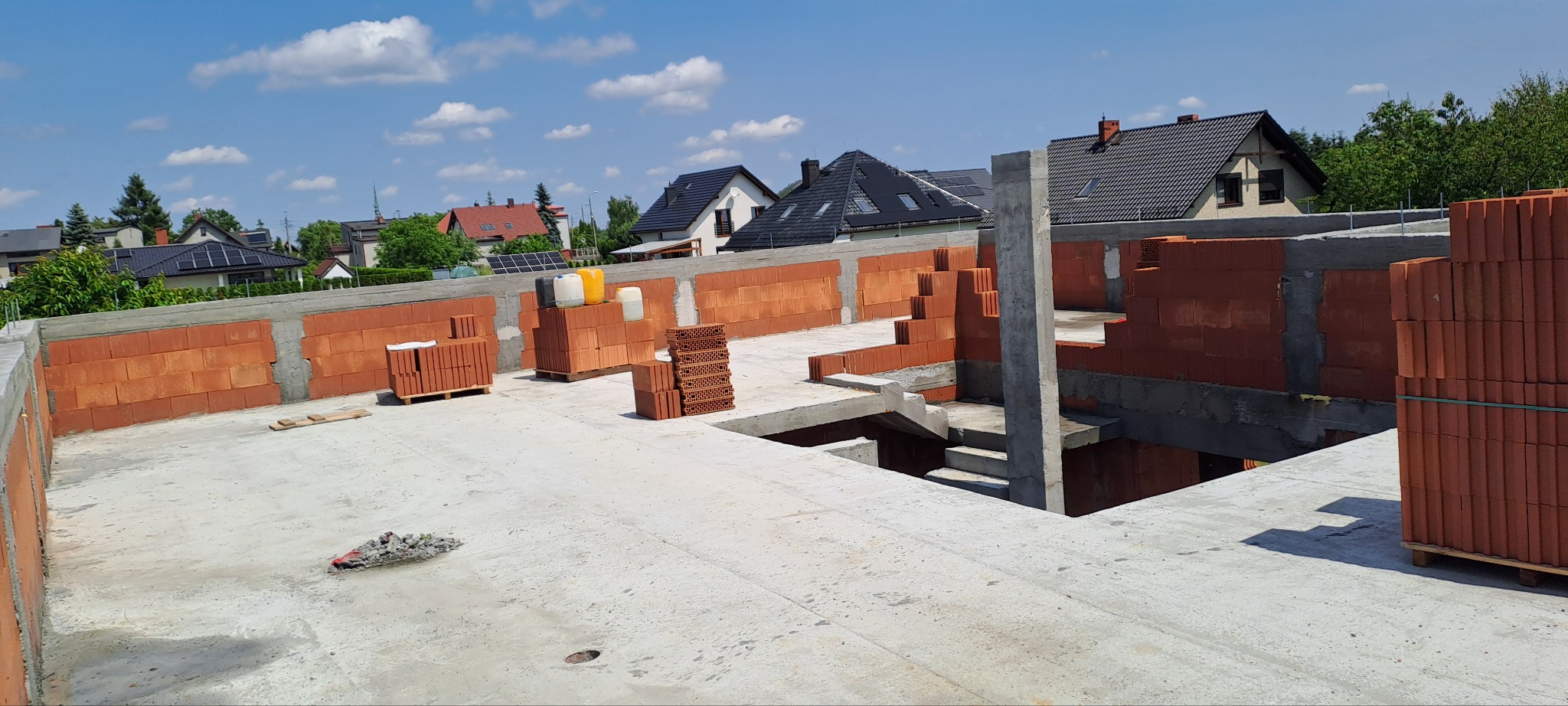 Budowa domu: widok z góry na betonowy strop z częściowo wymurowanymi ścianami z czerwonej cegły. W tle zabudowa jednorodzinna i błękitne niebo.