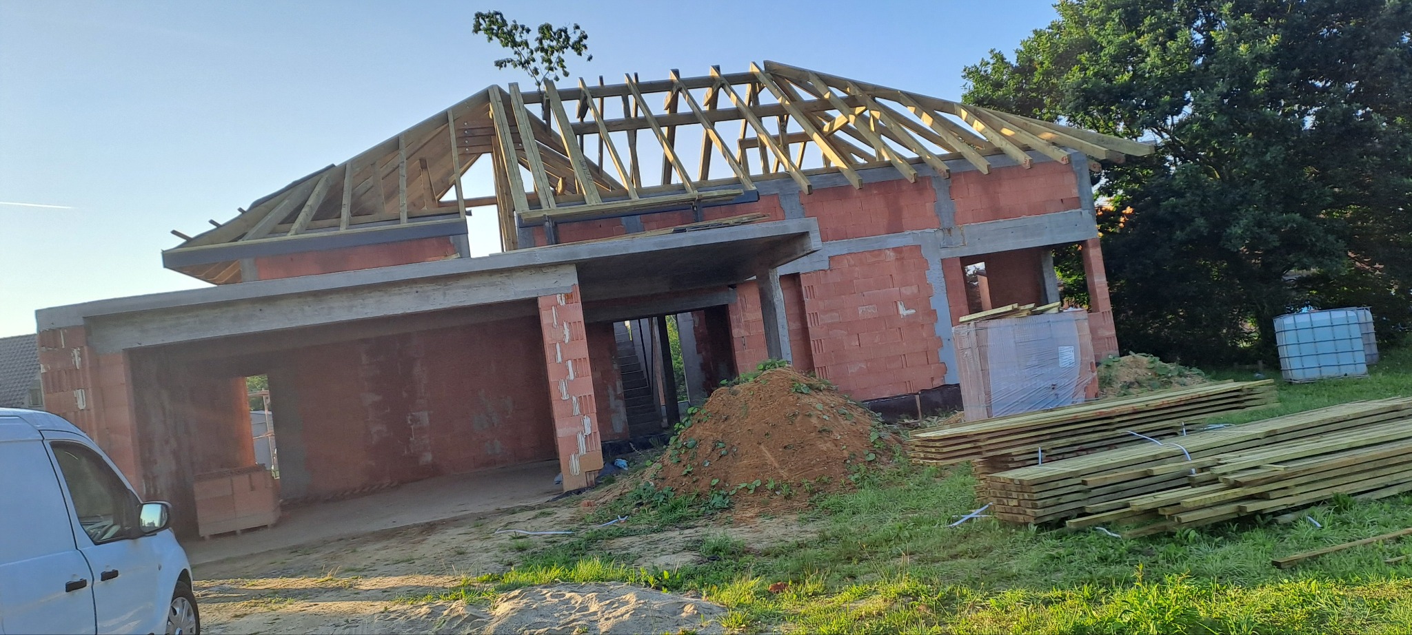 Surowy stan domu z czerwonej cegły z drewnianą konstrukcją dachu w trakcie budowy, widok z boku na tle zieleni i błękitnego nieba.