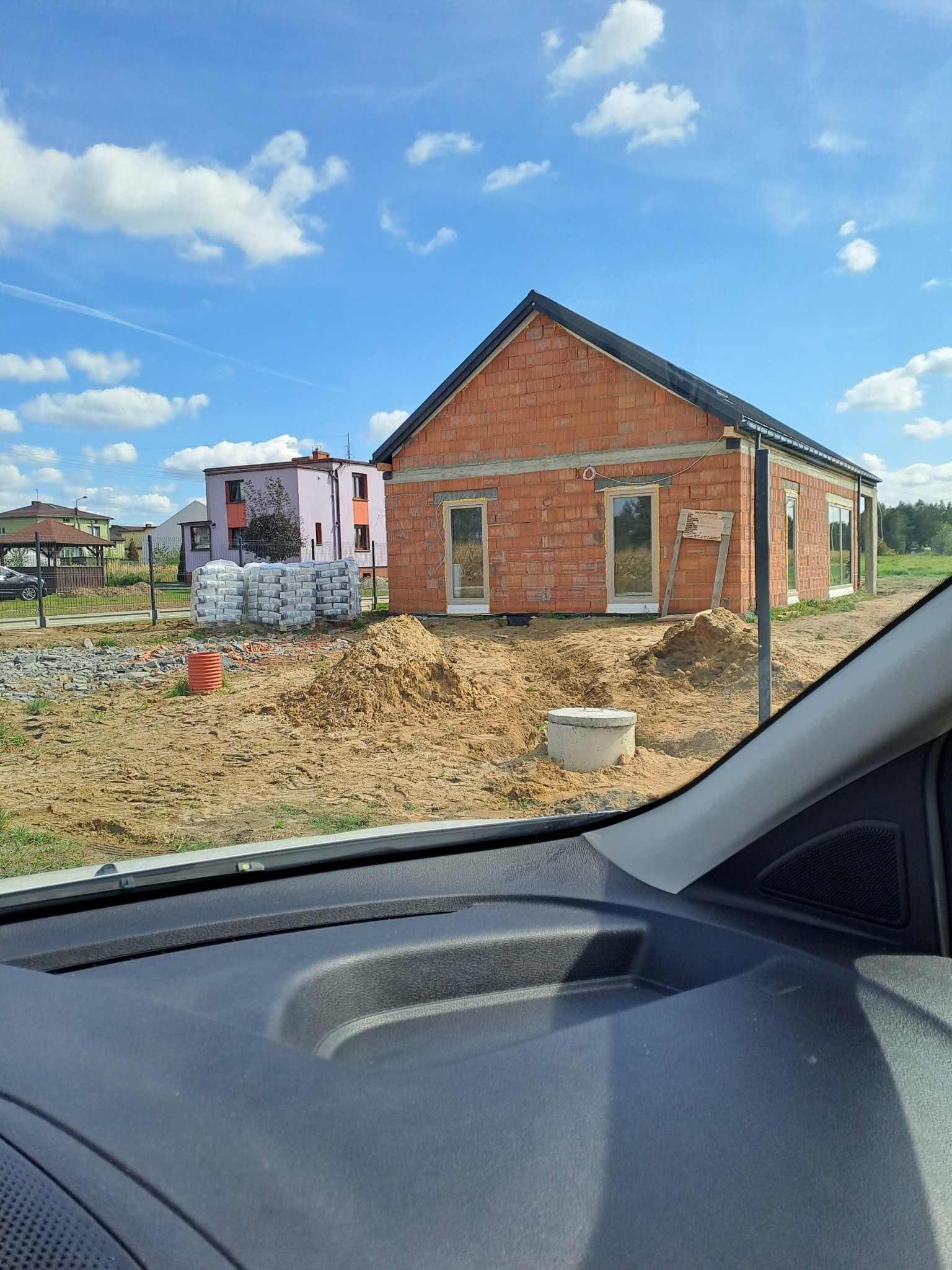 Budowa domu z czerwonej cegły w trakcie realizacji. Widoczne okna, sterta worków z zaprawą, piasek i fragment deski rozdzielczej samochodu.