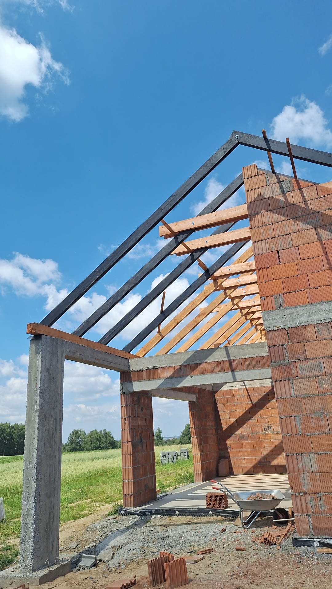 Wznoszenie domu z czerwonej cegły: widok na konstrukcję ścian, betonowy słup i drewniany szkielet dachu na tle błękitnego nieba z chmurami. Prace w toku, widoczna taczka.