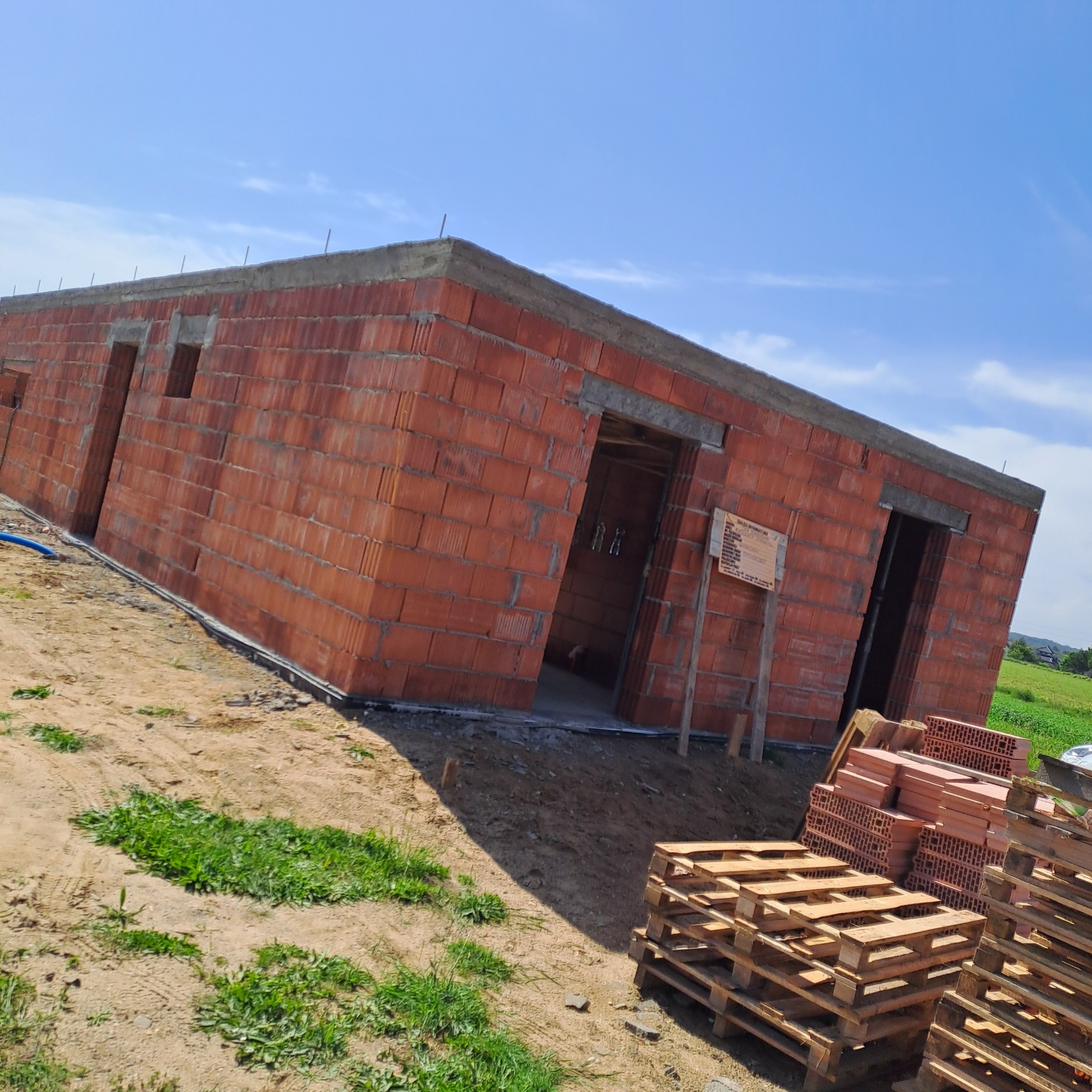 Niski budynek w trakcie murowania ścian z czerwonej cegły, widoczne otwory okienne i drzwiowe, palety z cegłami obok, tablica informacyjna budowy.