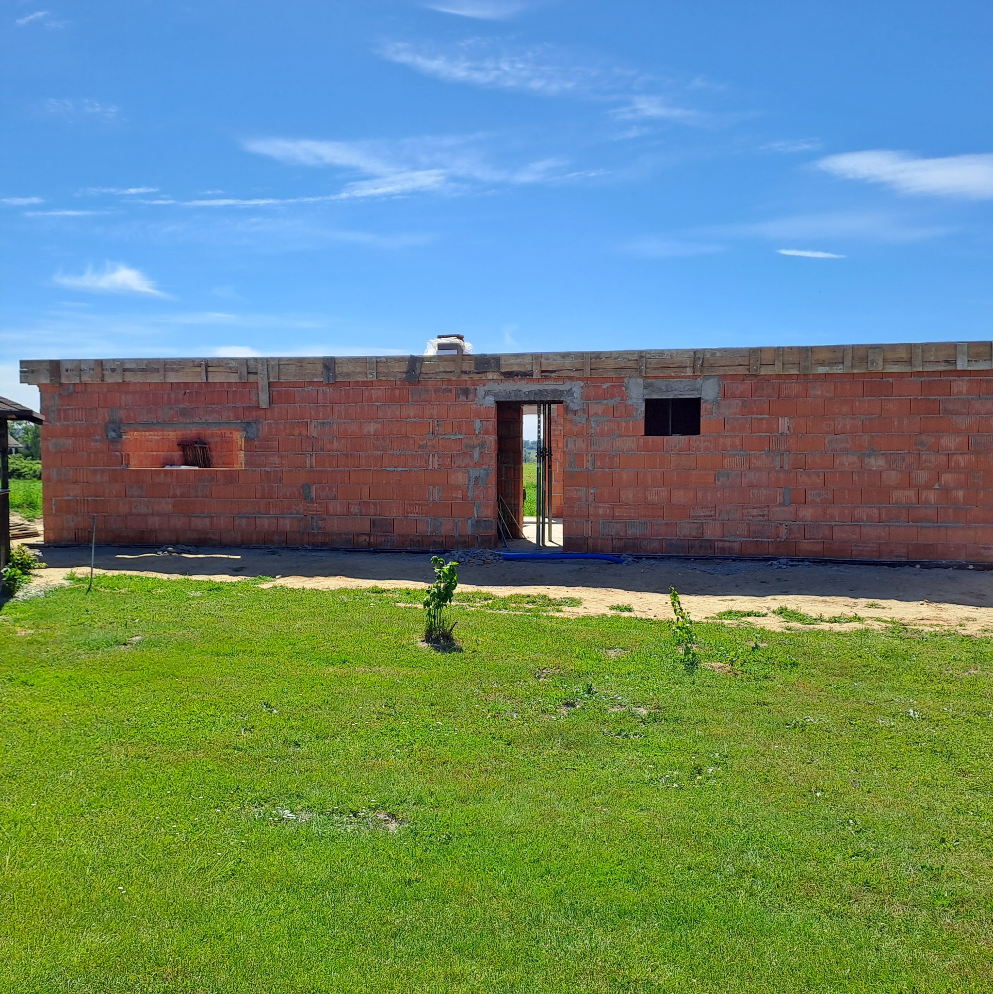 Niski budynek z czerwonej cegły w trakcie budowy, widoczne otwory okienne i drzwiowe. Drewniane elementy szalunku na górze. Zielony trawnik na pierwszym planie.