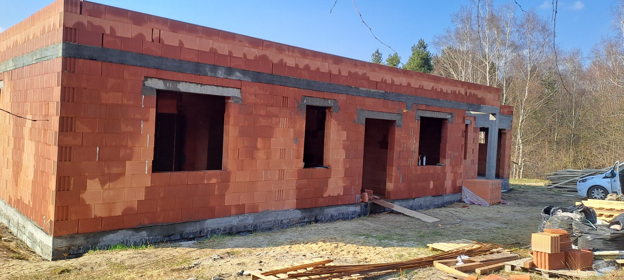 Surowy stan domu z czerwonej cegły, widoczne otwory okienne i drzwiowe, betonowe nadproża. W tle drzewa i częściowo widoczny samochód dostawczy.