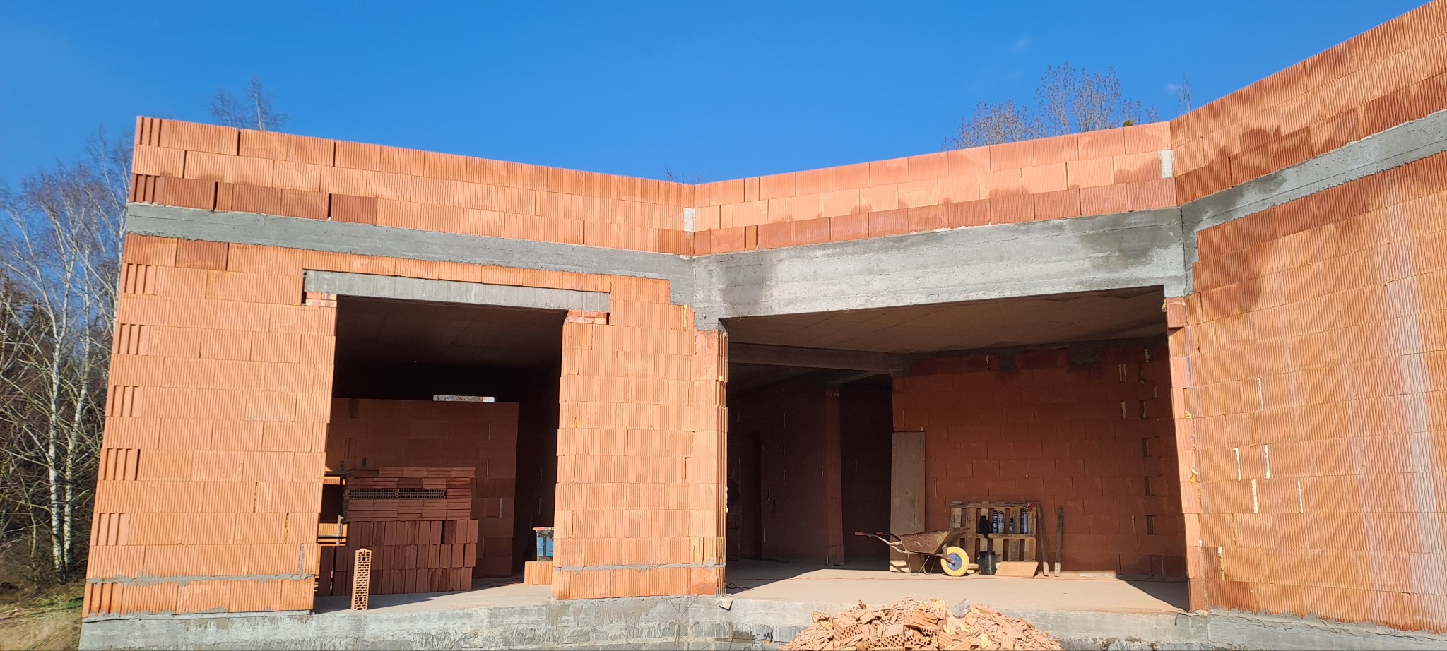 Wznoszenie ścian z czerwonej ceramiki poryzowanej, widoczne nadproża betonowe, w tle drzewa i błękitne niebo. Częściowo zrealizowany etap budowy domu.