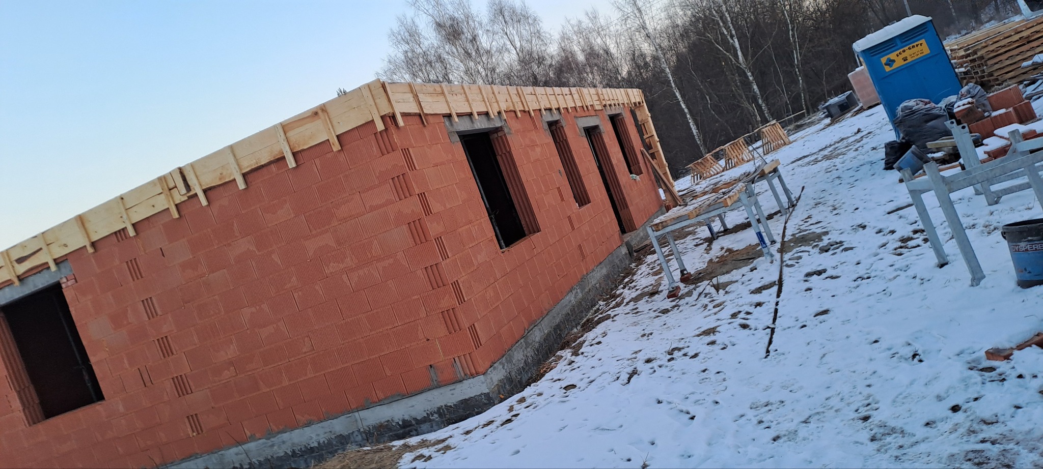 Ściana budynku w trakcie wznoszenia z czerwonej cegły dziurawki, z widoczną konstrukcją drewnianą u góry. Zimowy krajobraz z białym śniegiem.