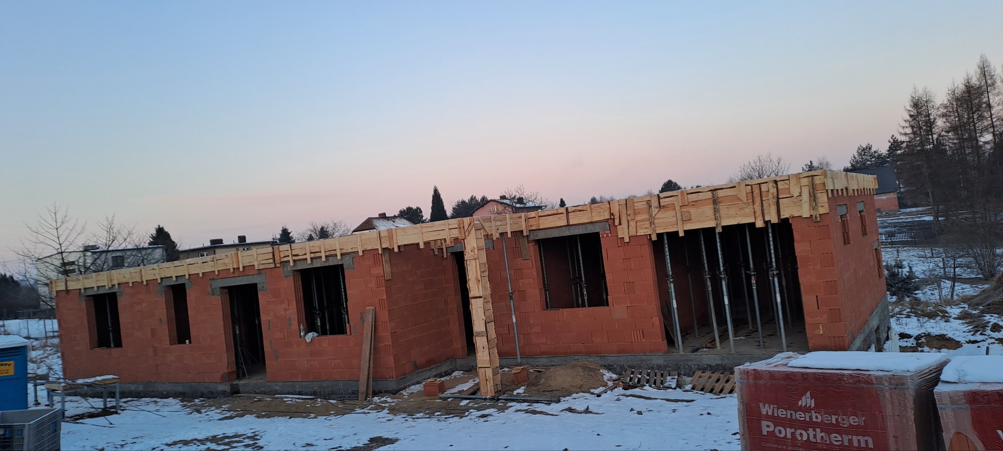 Wznoszenie ścian domu z czerwonej cegły Porotherm. Widoczne drewniane szalunki nad otworami okiennymi i podporami. Zimowy krajobraz w tle, paleta z cegłami Wienerberger.