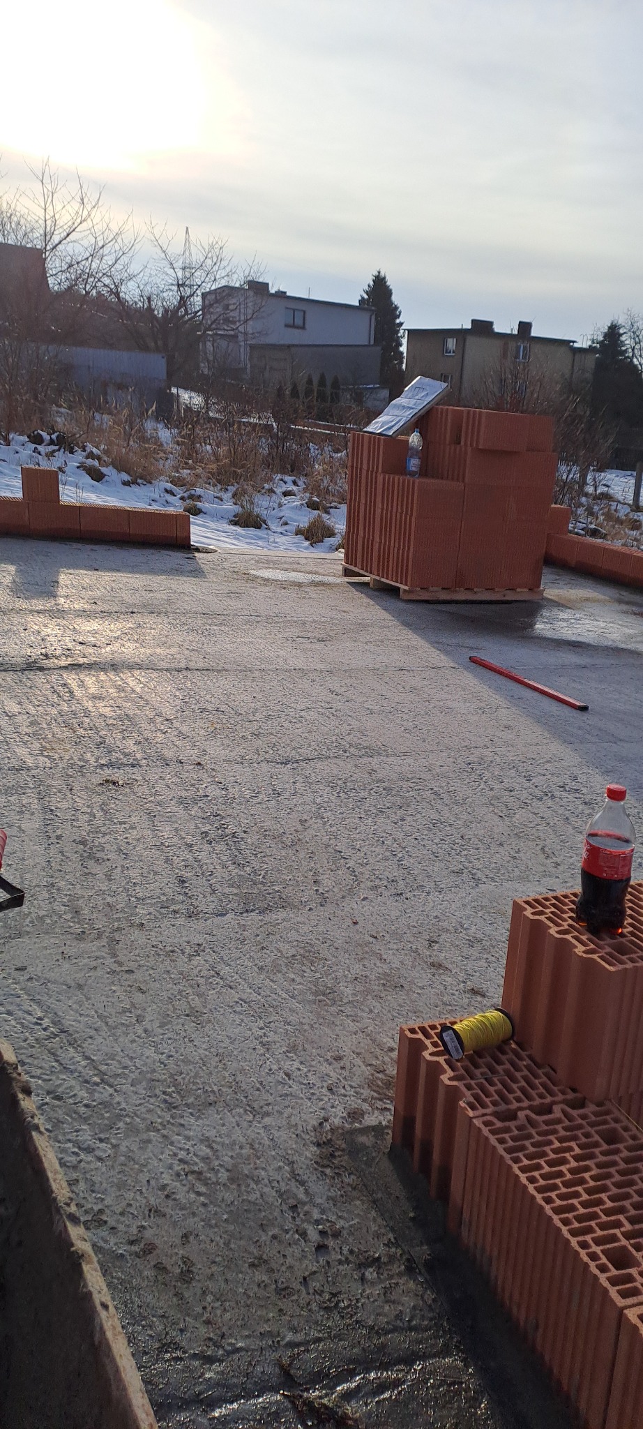 Świeżo wylana płyta fundamentowa z paletą cegieł ceramicznych i butelką napoju. Widoczne narzędzia murarskie i zabudowa w tle. Słoneczny dzień.