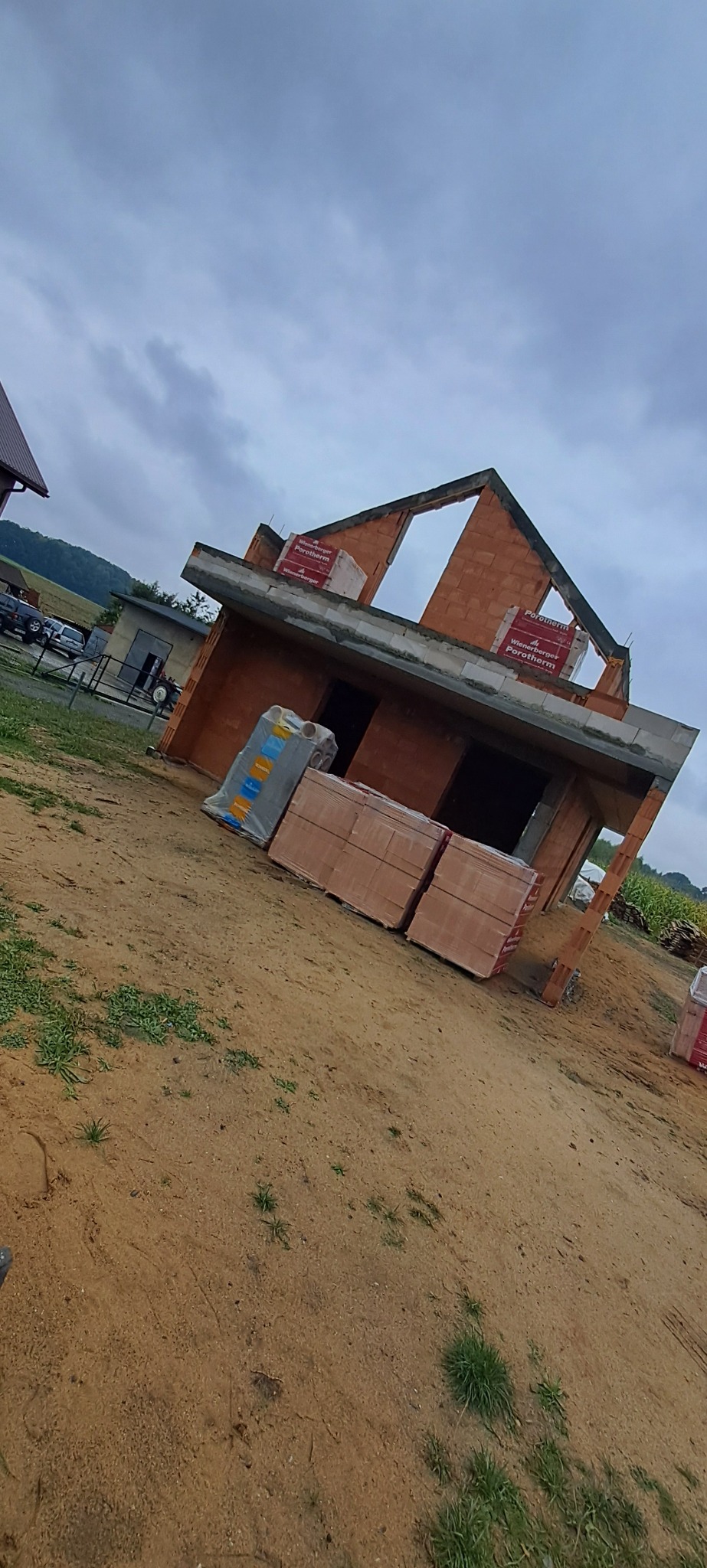 Niedokończony, ceglany dom w stanie surowym otwartym z paletami cegieł na placu budowy, widok z dołu, pochmurne niebo w tle.