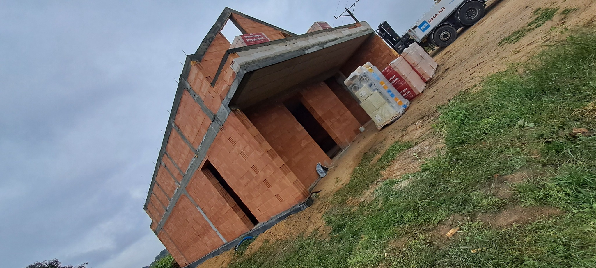 Wznoszenie dwukondygnacyjnego domu z cegły ceramicznej Porotherm na tle pochmurnego nieba, obok zaparkowany samochód dostawczy z materiałami budowlanymi.