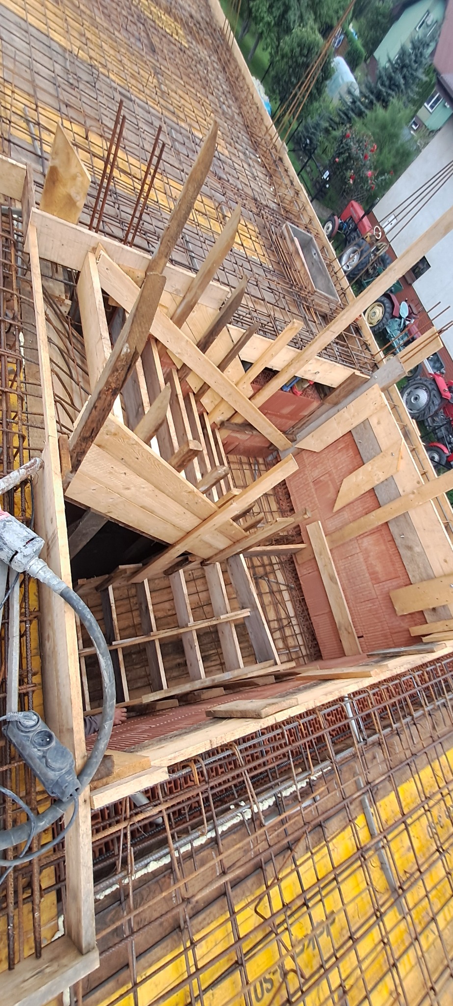 Zbrojenie pod fundamenty z drewnianym szalunkiem, widok z góry. Na drugim planie budynki i dwa czerwone traktory. Elementy zbrojenia częściowo poza ujęciem.