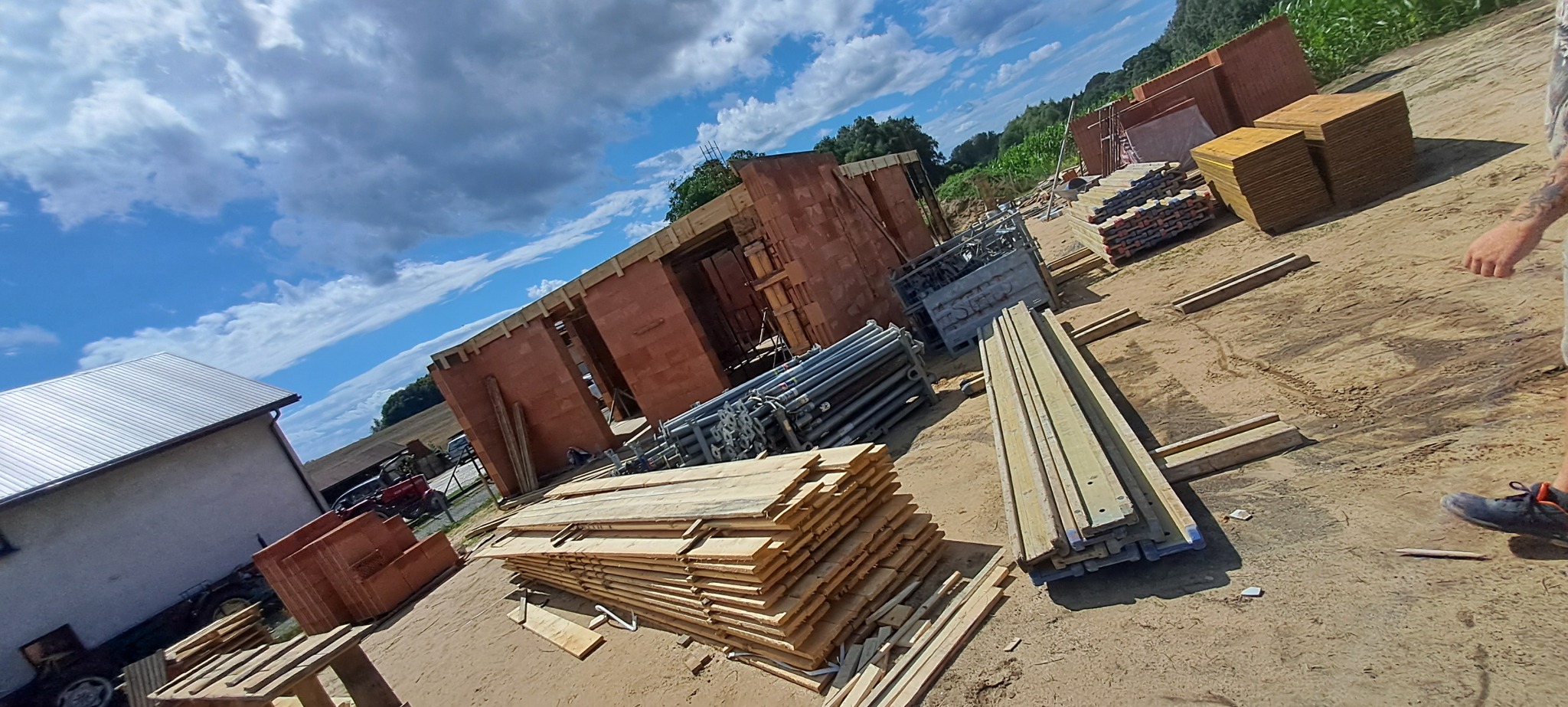 Budowa domu w toku: wzniesione ściany z czerwonej cegły, drewniane deski, podpory i szalunki na placu budowy z błękitnym niebem w tle.