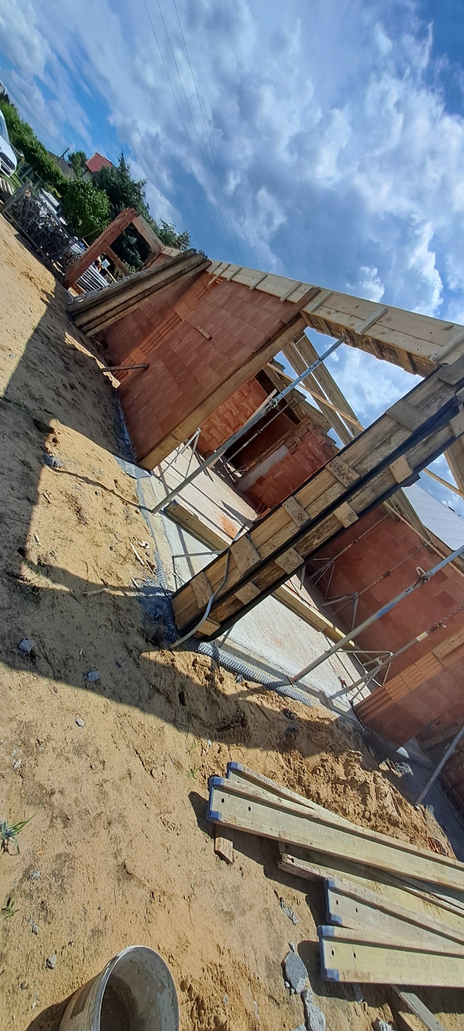 Budowa domu z czerwonej cegły, widok z dołu na konstrukcję i niebo w Pszowie. Widoczne drewniane elementy szalunkowe i metalowe podpory.