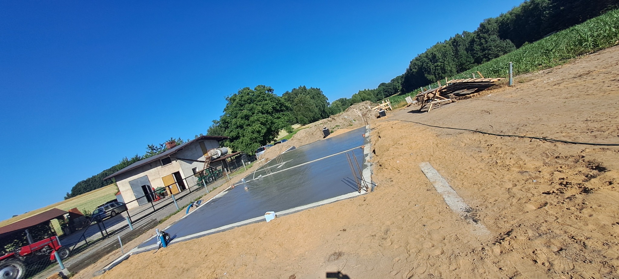 Świeżo wylana, gładka płyta fundamentowa na budowie w słoneczny dzień. Widoczna zbrojenia, otoczenie placu budowy, częściowo dom i drzewa w tle.