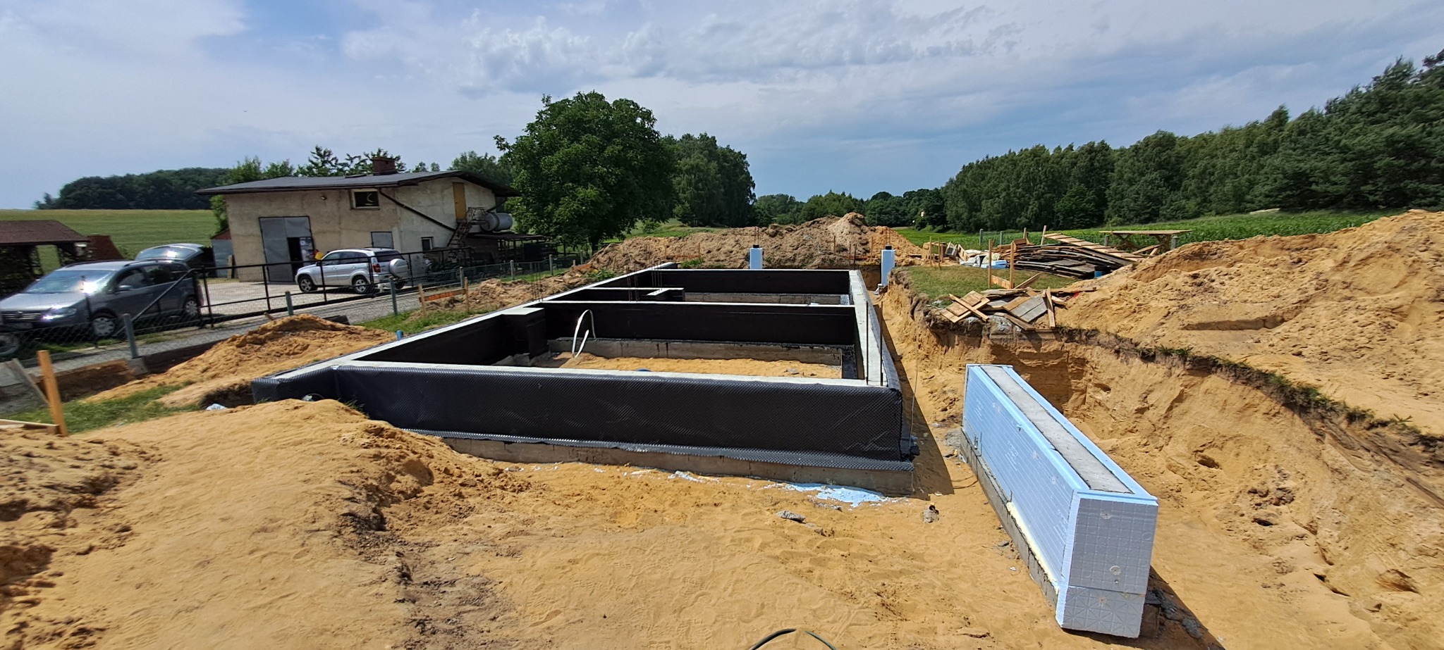 Przygotowany zarys fundamentów budynku z hydroizolacją i ociepleniem, widok z góry na wykop z piaskiem, budynek gospodarczy i samochody w tle.