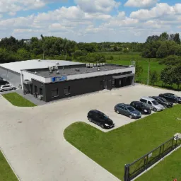 Zdjęcie z lotu ptaka przedstawia nowoczesny budynek hali z biurem, z szarymi ścianami, białym dachem i czarną elewacją biurową, zaparkowane samochody osobowe, teren zielony i błękitne niebo...