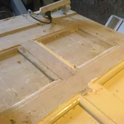 Drewniane drzwi z widocznymi śladami szlifowania i przygotowania do malowania, z brązowym pędzlem w lewym dolnym rogu i przedłużaczem elektrycznym w górnym rogu.