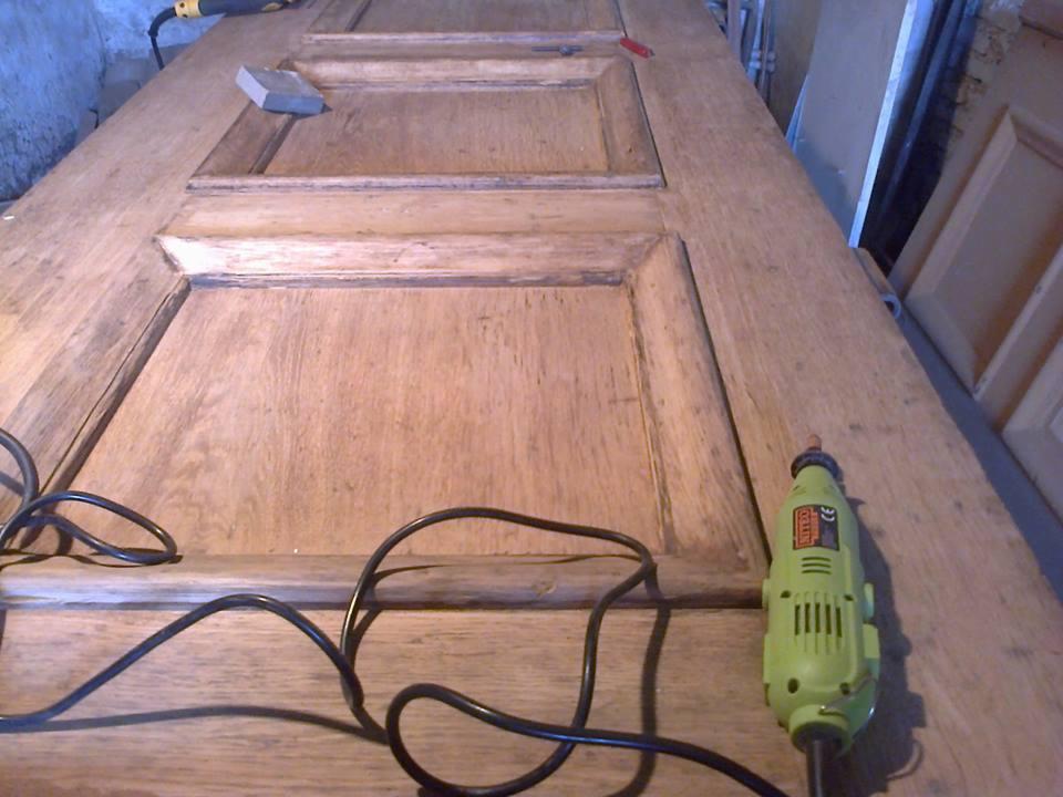 Drewniane drzwi z panelami w trakcie renowacji, szlifowane szlifierką oscylacyjną i narzędziem wielofunkcyjnym, widoczne ślady starej farby i przygotowania do malowania.