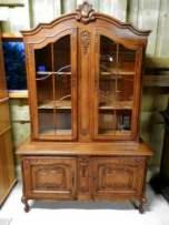 Antyczny, dębowy kredens z przeszkloną nadstawką, bogato zdobiony rzeźbieniami, ustawiony w słabo oświetlonym pomieszczeniu.