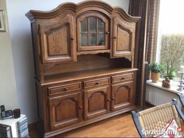 Rustykalna, dwuczęściowa komoda z litego drewna dębowego z trzema szufladami i przeszkloną szafką górną, ustawiona w salonie obok okna i plecione krzesła.